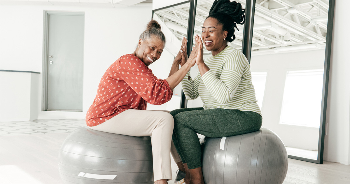 Two Black women, one older and one younger, seated on exercise balls and high-fiving with both hands.