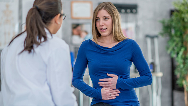A young woman in a blue sweater holding her hands over her stomach while speaking with a health care provider in a hallway.