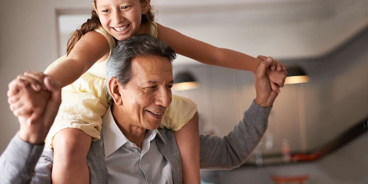 A man in a gray shirt standing indoors with his daughter seated on his shoulders.