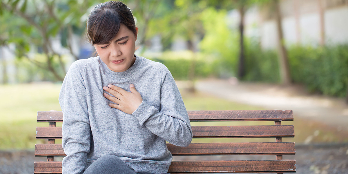 Young woman sitting on a bench with her eyes closed, clutching her chest in discomfort.