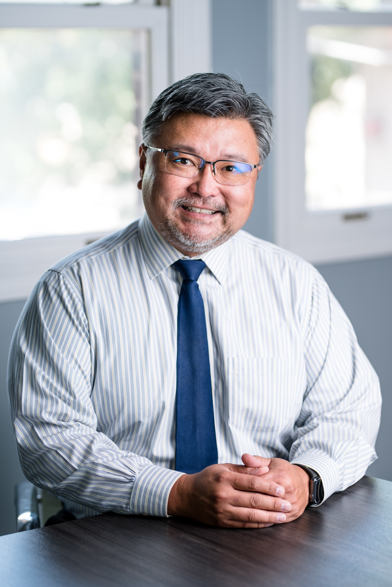 Pediatric cardiologist Frank Wong, MD, smiling while seated at a conference table in front of a window, wearing a white shirt and blue tie