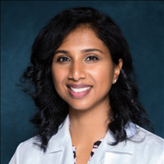 Dr. Jeny Ghartey wearing a white coat and smiling in front of a dark blue backdrop.