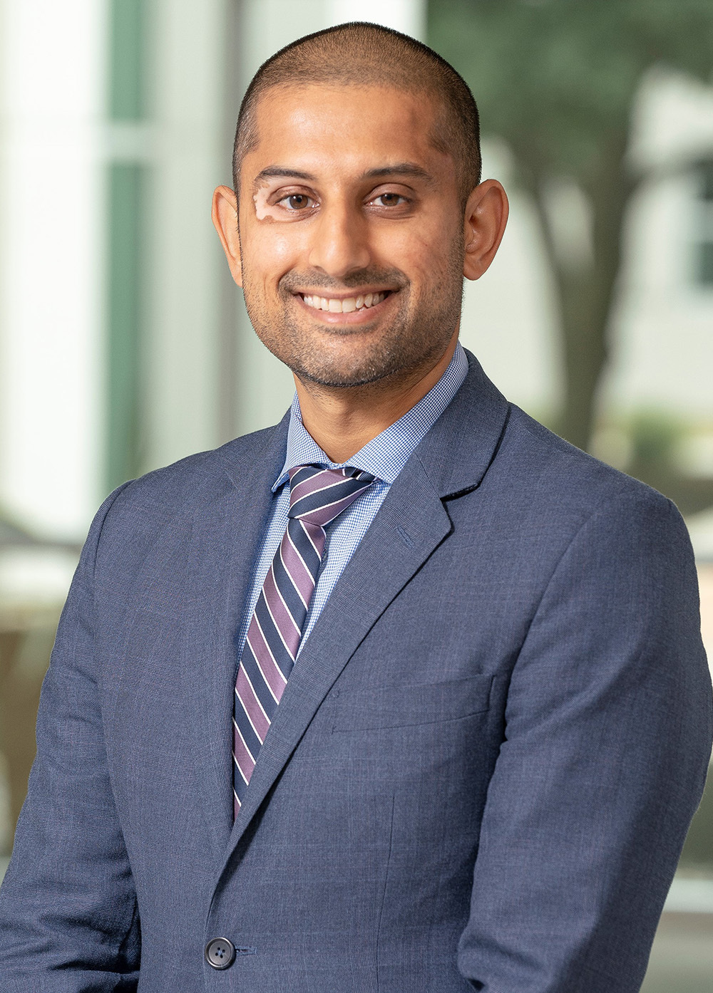 Mark Gajjar in a gray suit and striped tie and smiling in front of a tree and a column of windows.