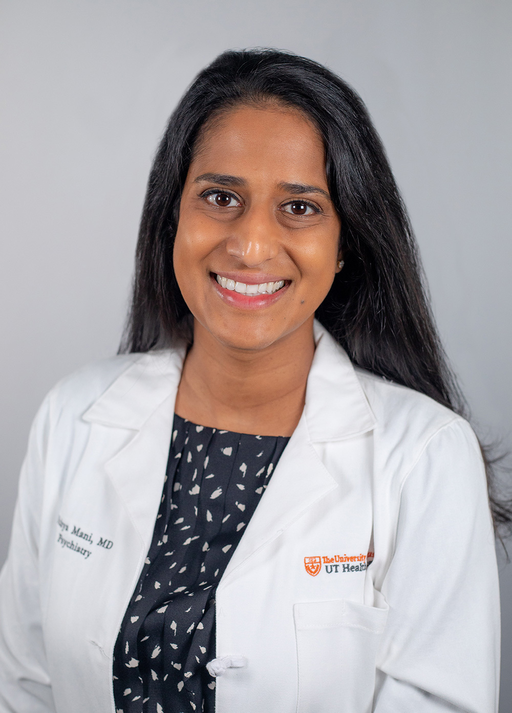 Dr. Nithya Mani wearing a white coat and smiling in front of a white backdrop.