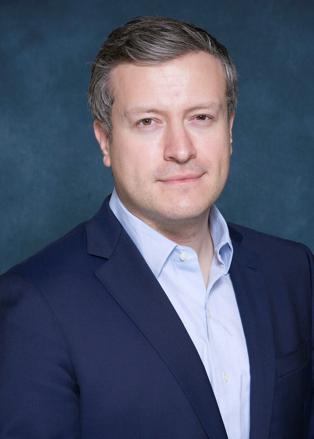 Dr. Peter Monteleone wearing a blue suit and smiling in front of a dark blue backdrop.