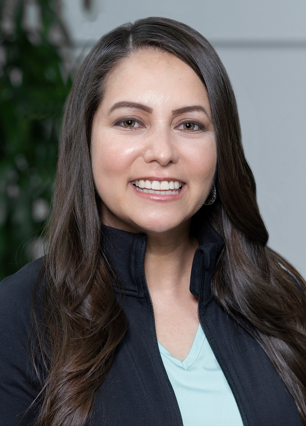 Vanessa O'Dell, social worker stands in front of a plant and white wall in a dark suit smiling.
