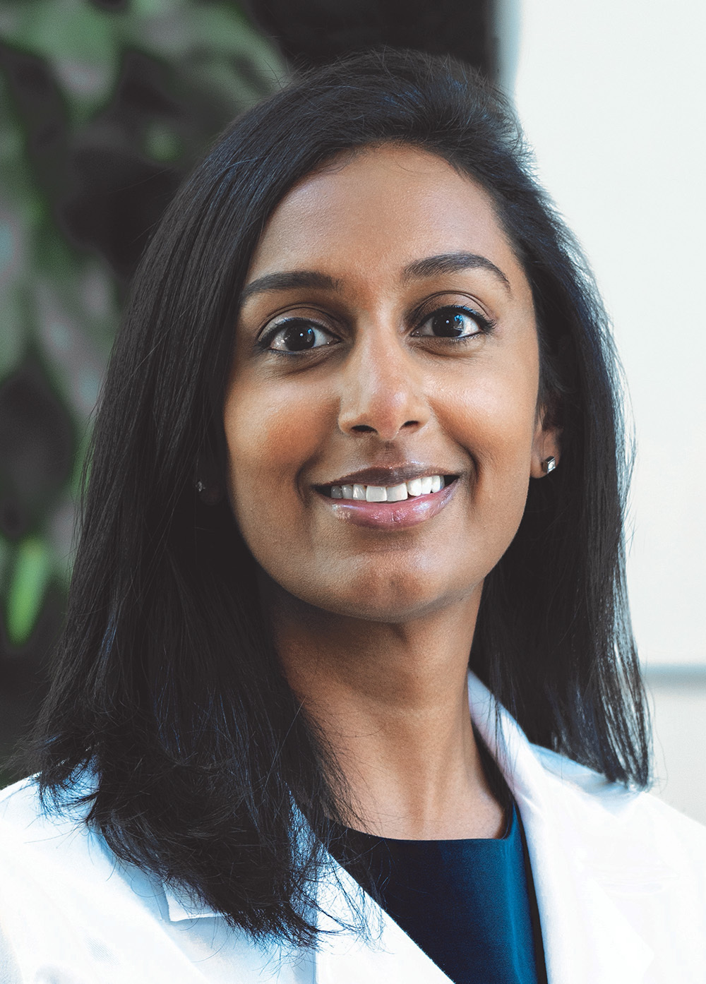 Dr. Sneha Vakamudi in a white coat and smiling in front of a wall and greenery.