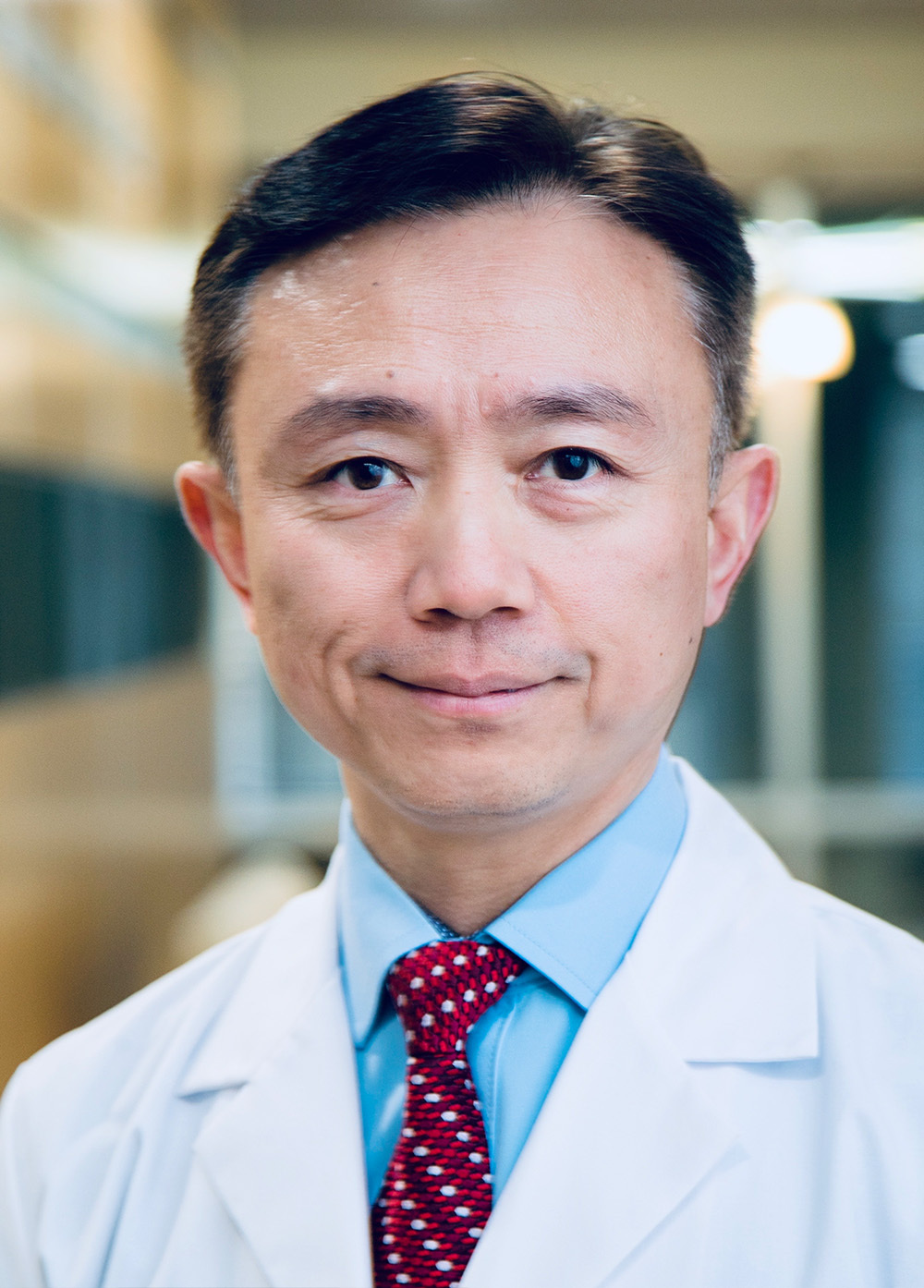 Dr. Yan Liu wearing a white coat and red necktie and smiling indoors.