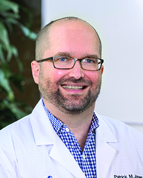 Dr. Patrick Jones wearing a white coat and smiling near a tree and a white wall.