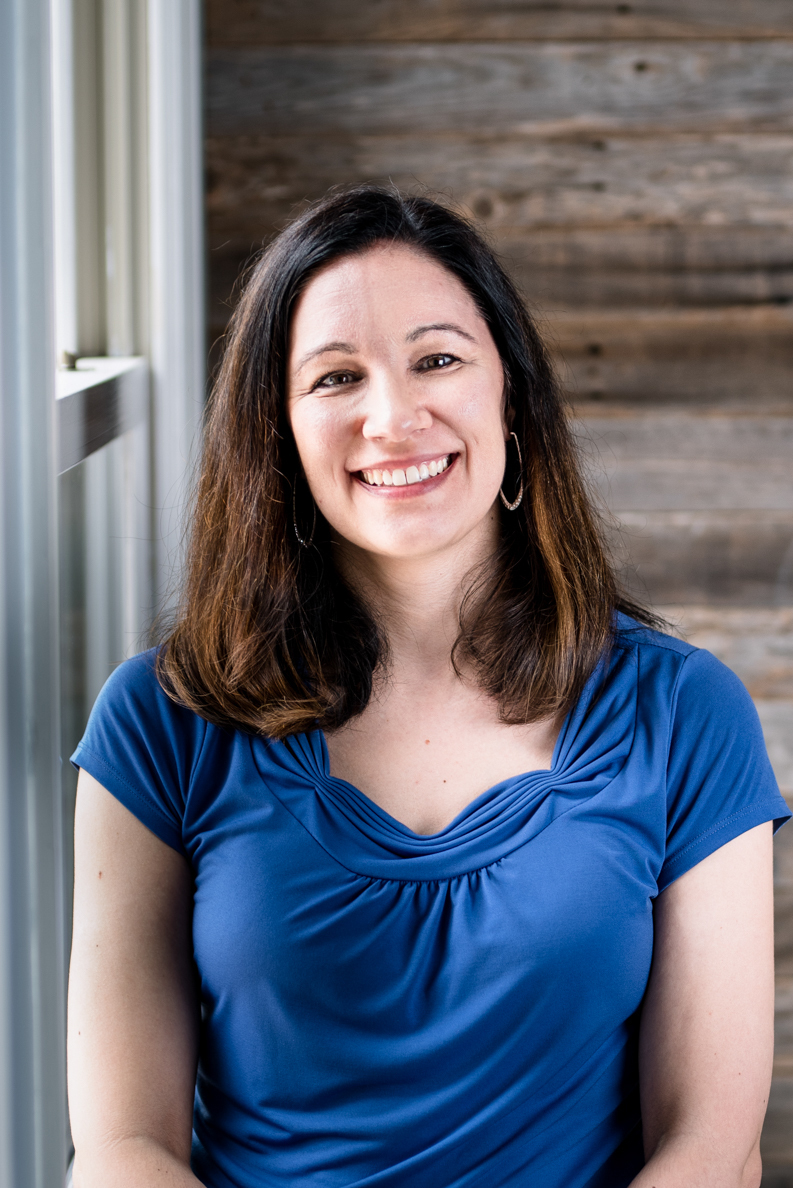 Christine Dillingham wearing a blue blouse and smiling in front of a wooden wall.
