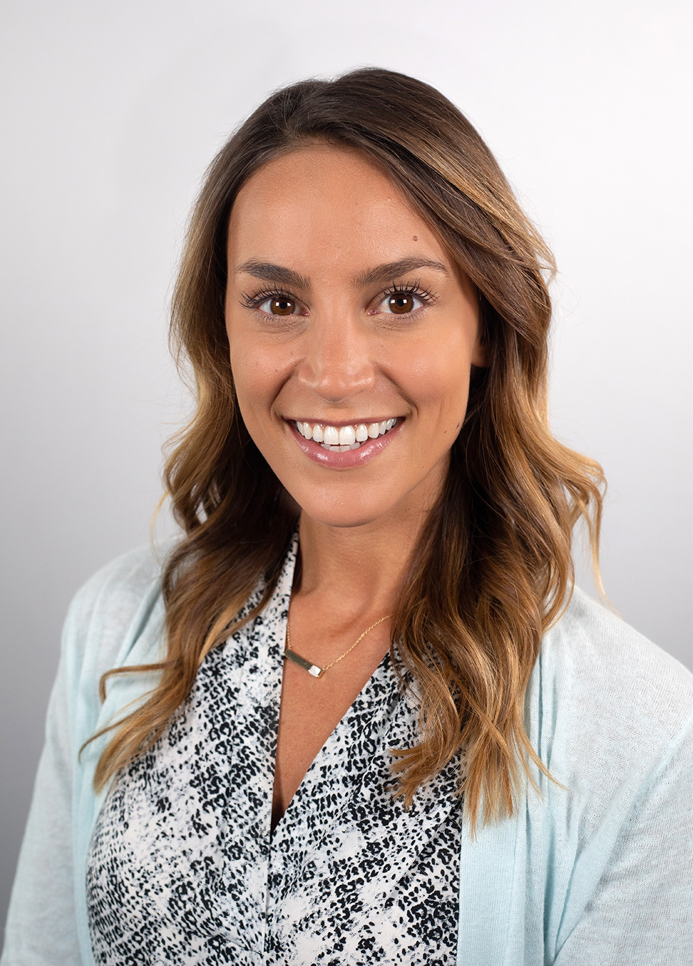 Clara Di Vincenzo wearing a blue cardigan and patterned blouse and smiling in front of a white backdrop.