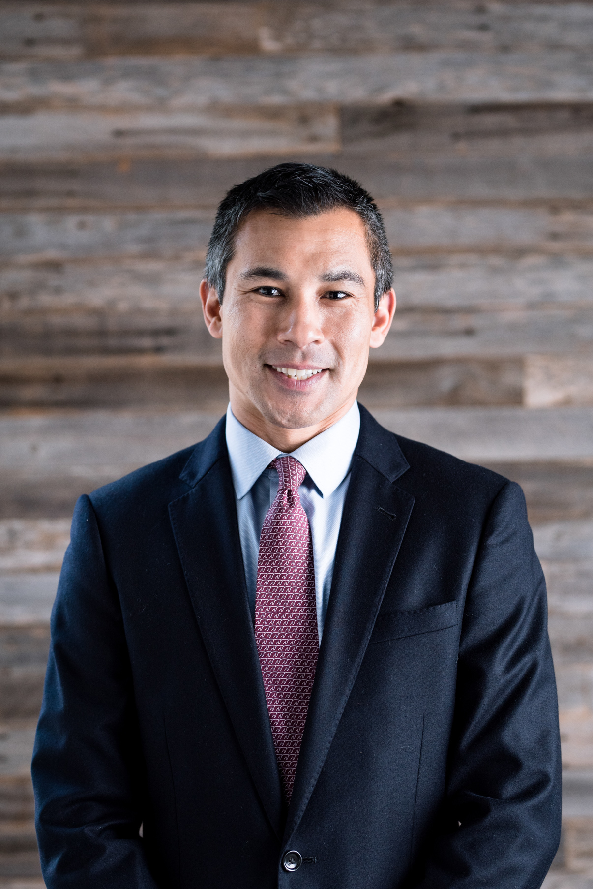 Pediatric cardiologist Joseph Casadonte, MD, wearing a suit and red tie and smiling in front of a wooden wall.