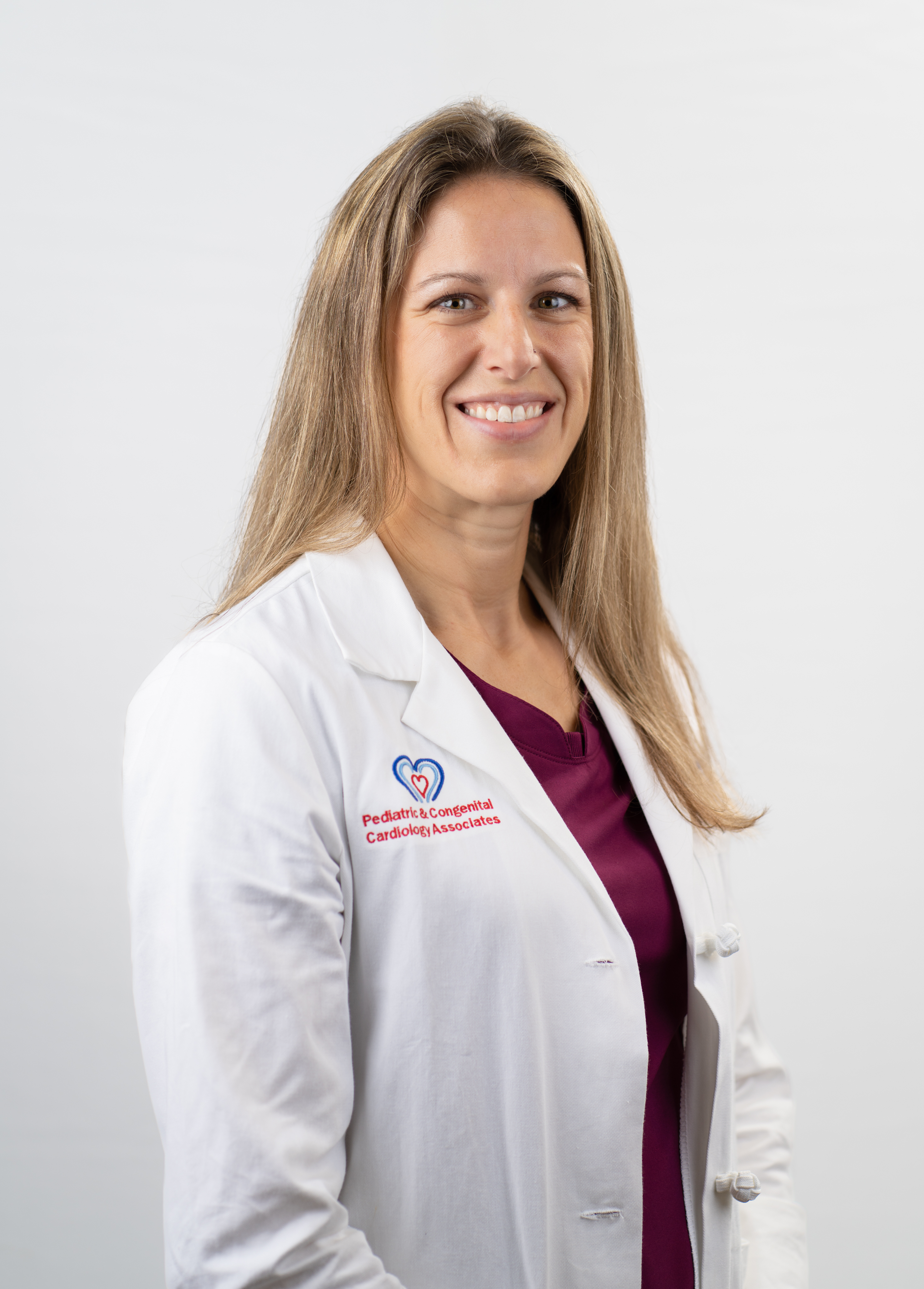 Acute care pediatric nurse practitioner Katie Aldrige, CPNP, APRN, smiling in front of a white backdrop. She is wearing a white coat with an embroidered Pediatric and Congenital Cardiology Associates of Texas logo.