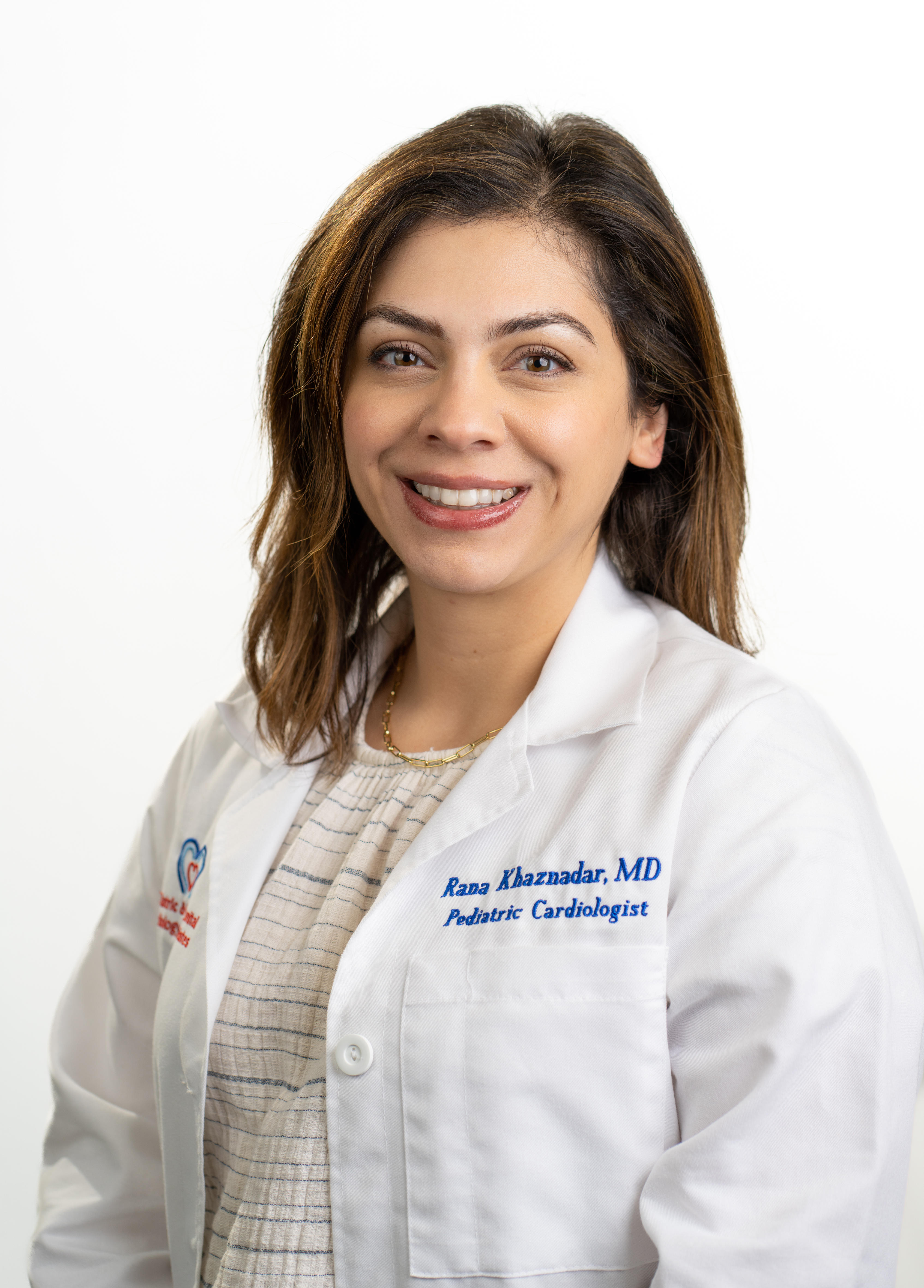 Pediatric cardiologist Rana Khaznadar, MD, smiling in front of a white backdrop. She is wearing a white coat with an embroidered UT Health Austin logo.