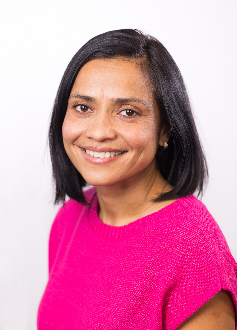 Genetic counselor Raynah Lobo, MS, LCGC, wearing a pink blouse and smiling in front of a white backdrop.