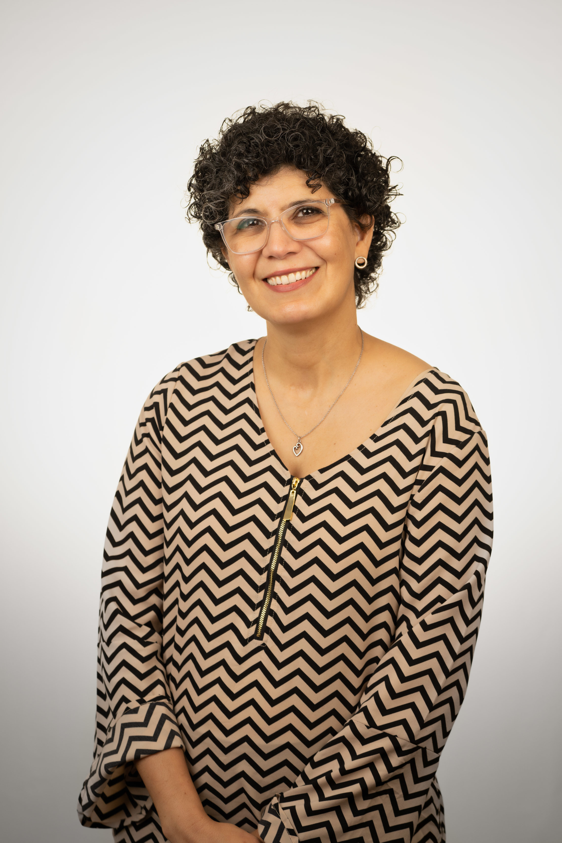 Maira Souza wearing a chevron patterned blouse and smiling in front of a white backdrop.