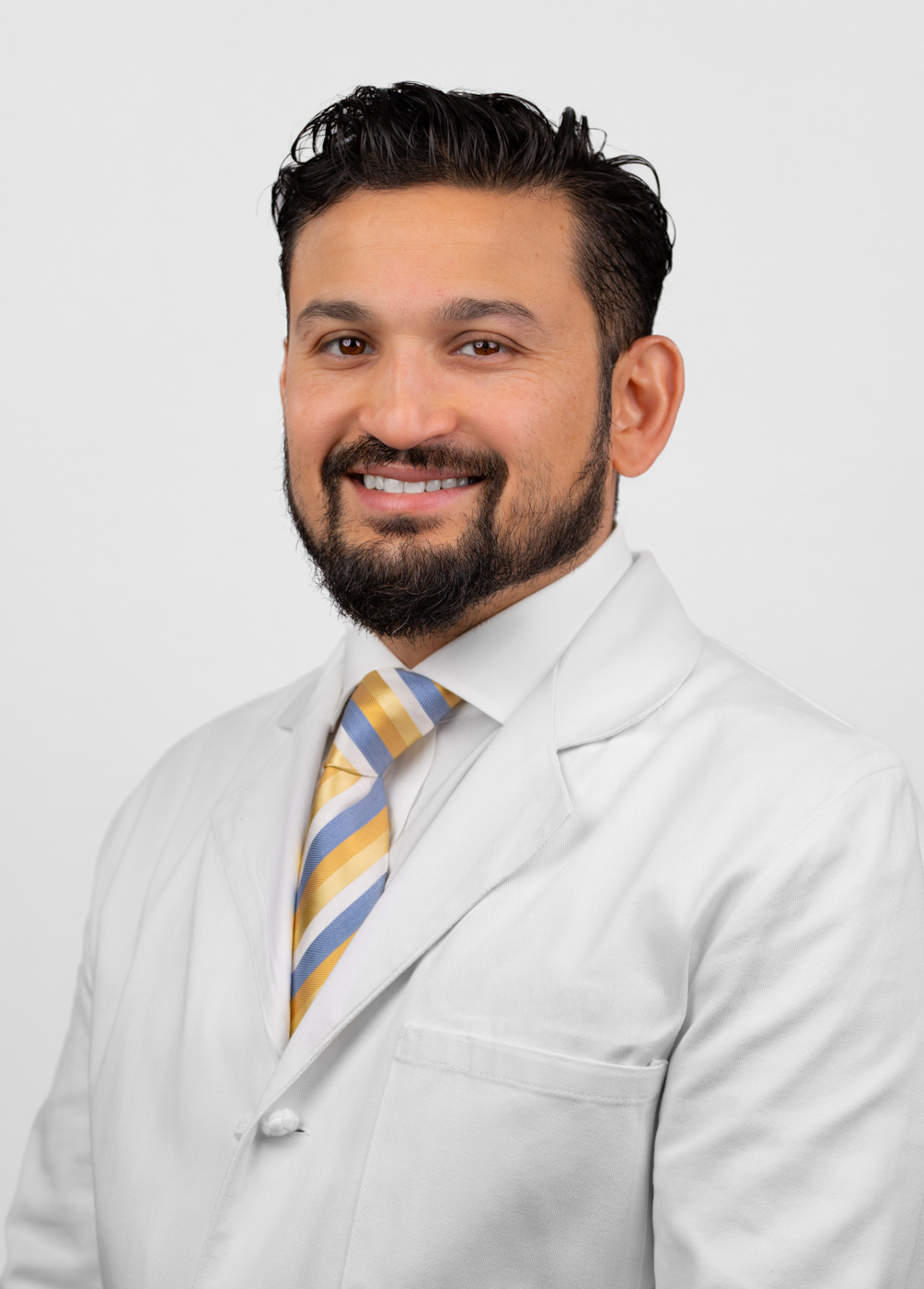 Headshot of Omar Iqbal. He is wearing a white coat, white dress shirt, and a yellow and blue tie.